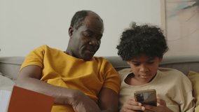 African American father and son using smartphone sitting on sofa