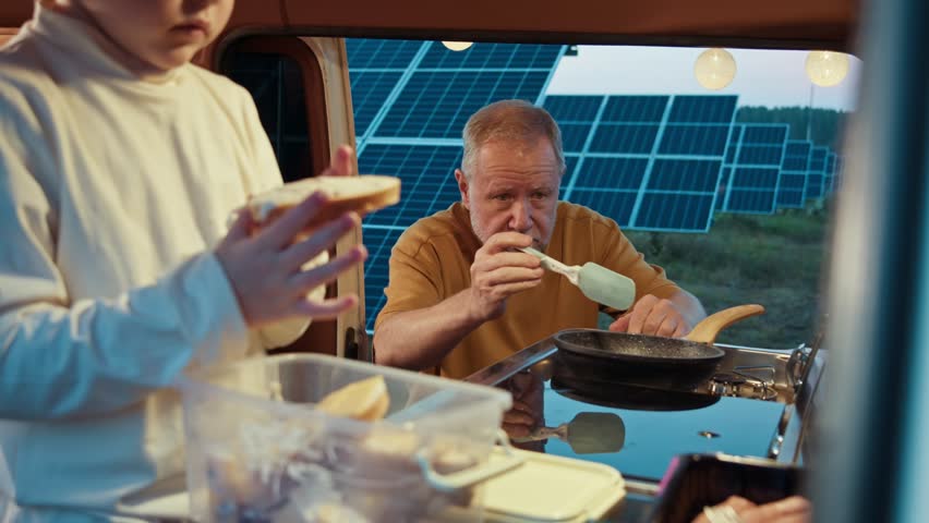 A boy eats a sandwich in a camper as the man cooks beside him, both enjoying the evening at the solar farm