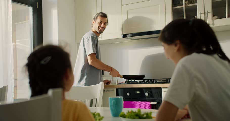 A young brunette man shows how he can throw toast in a frying pan for his wife and little daughter while preparing breakfast in the kitchen. The man s wife and daughter watch him prepare the morning Royalty-Free Stock Footage #1112118649