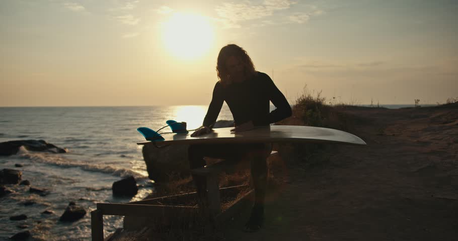 A young guy with curly hair in a black wetsuit sits on a bench near the sea holds a surf board in his hands and cleans it and sets it up for surfing at sunrise in the morning in summer