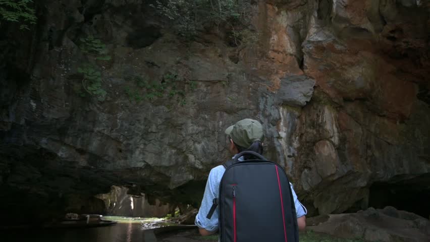 Rear view of female tourist with camera bag is walking into the cave that water stream flowing inside under the huge rocky mountain. Outdoor pursuit and Leisure activities. Solo travel.