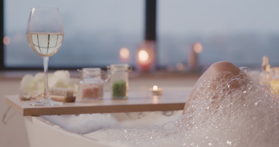 Camera focuses on a wooden board above a bathtub with a glass of wine, bath salts and a candle. Royalty-Free Stock Footage #1112127623
