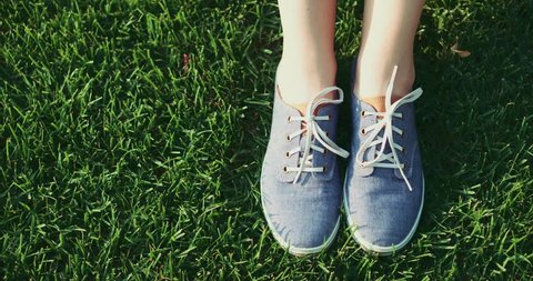 Female feet in sneakers gumshoes moving and posing on a sunlit green grass, close up. Woman's feet in blue jeans color sports shoes. Slow motion, 120fps. 4K, DCi.