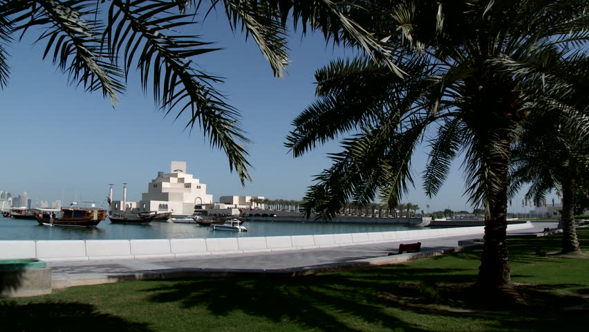 A pan between date trees of the city and waters in Doha, Qatar showing traditional Arabian wooden boats docked on the water and the city skyscrapers in the background