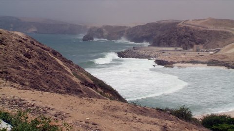 A pan showing the rocky, mountainous coast line of Salalah, Oman as the sea comes into a sandy white beach and waves crash into rocks in the distance