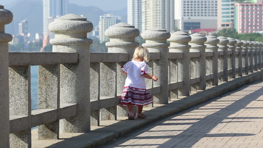 little blonde girl in Ukrainian national blouse runs by barrier on embankment