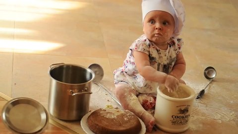 A little cute ten-months-baby-girl with cooking hoods on her head is sitting on the kitchen floor, she is heavily soiled with wheat flour - she's playing cook