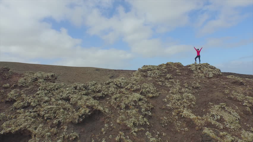 AERIAL: Flying around cheerful young woman standing on the edge of big volcano crater with hands raised