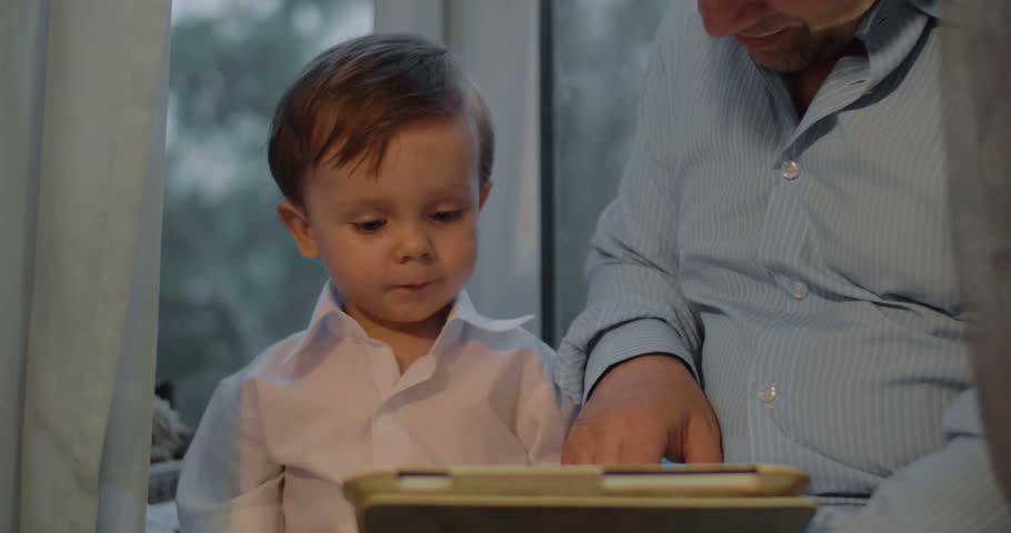 Little boy is sitting by his father and watching something in tablet PC.