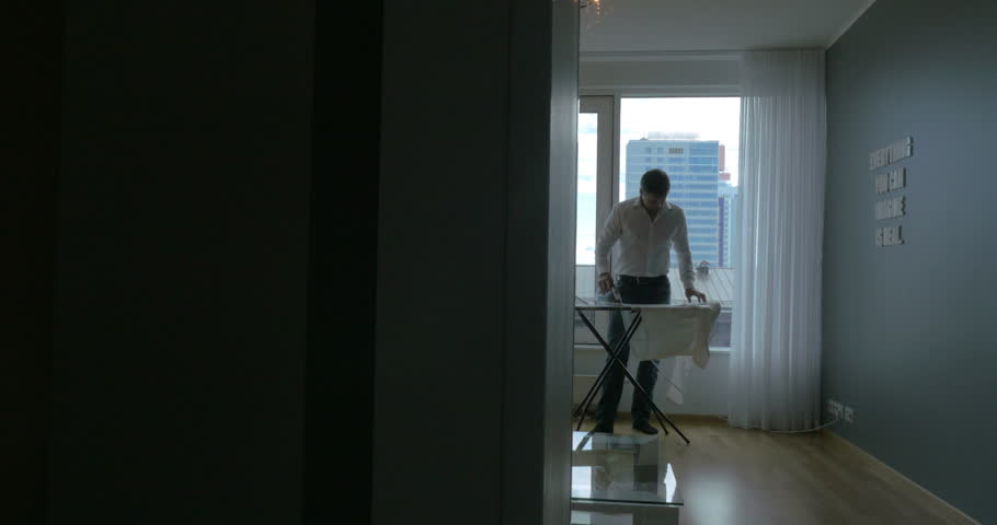 Steadicam shot of a youg man ironing the clothes at home. He standing by the big window with city panorama