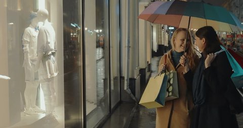 Two female friends are standing in front of the show window of clothing shop and talking. They are holding shopping bags in hands.