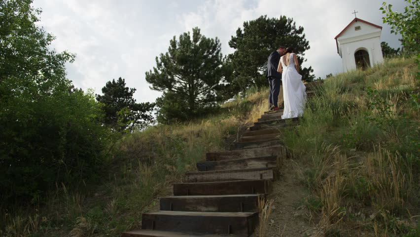 A newly married couple climbs a staircase to a shrine with a cross above.  4k. In slow motion.
