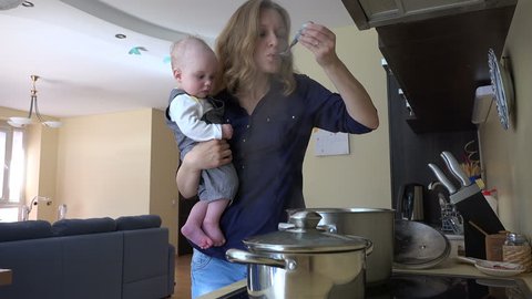 Nanny with infant baby in hands cooking mix meal dish in pot and taste it with spoon. Housewife multitasking at home. Vapor steam rise. Static shot. 4K UHD video clip.