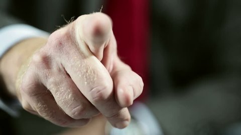 Businessman's hand comes into focus, lifting from his lap and pointing at the camera, indicating or accusing.