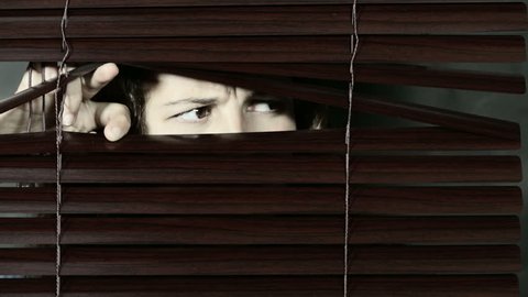 Young woman opens dark Venetian blinds and peeps through them, frowning, looking nervously from side to side, then to camera and shaking her head. Copy space on closed blinds.