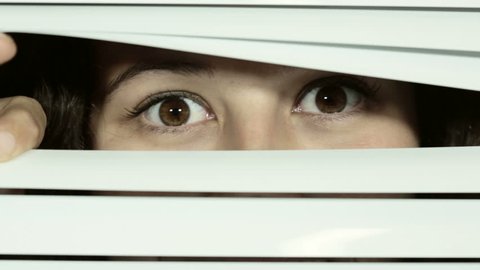 Pretty brown-eyed girl looks through white Venetian blinds at camera, smiles in happy recognition, then raises her eyebrows looking both knowing and flirtatious.