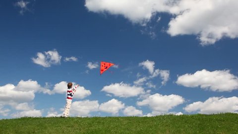 boy plays with orange kite in meadow against blue sky with white cloud