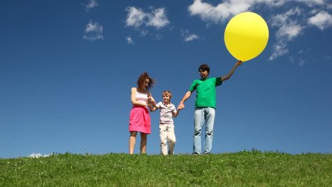 family of three holding yellow balloon hands down hill with green grass and wildflowers 
