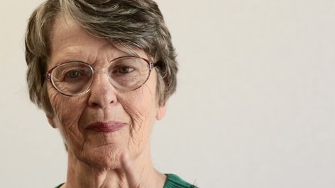 Pan across pale background to old woman's face. She looks at camera, hears or sees something, smiles, and nods in agreement, her hand on her chin. Copy space on the background.