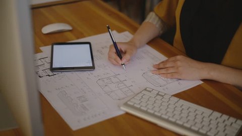 Young female architect working in office with blueprint signing details and making changes to the drawings. On the table lay a tablet, mouse and keyboard. on a video, we see a woman in modern clothes