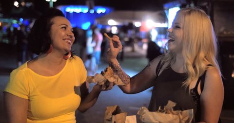 4K A hip trendy woman jokes around and offers friend a bite of her delicious street food truck french fries in a urban outdoor music festival night setting.