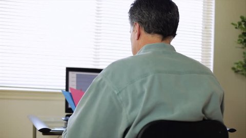A man sitting at a computer starts rubbing his neck and shoulder as if trying to relieve pain. Camera dolly shot
