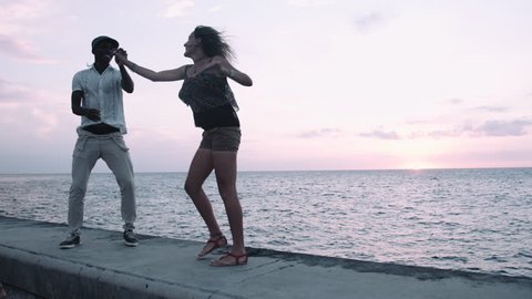 Young man and woman enjoying salsa dance on retaining wall at sunset, sea in background