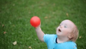 Video baby taking its first steps. Child with a color ball goes over the grass lawn.