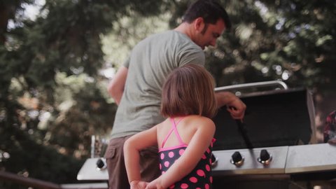 A little girl watches her dad clean a barbecue