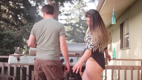 A husband and wife on their porch barbecuing and petting their dog