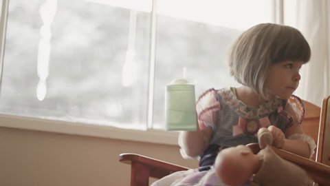 A little girl wearing a princess dress sits by a window and drinks from a sippy cup