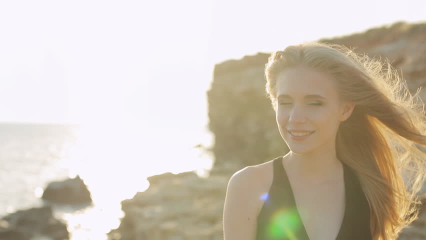 Beautiful blonde with long hair wearing black dress laughing near the sea at sunset in the mountains