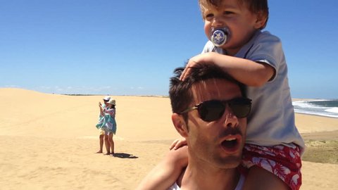 Child on top of dad's shoulders. 5 year old kid standing on father's shoulders giving support. Family in standing outside at the beach wearing sunglasses and having fun during summer holiday vacations