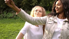 Two friends, young pretty girls, caucasian and african, having fun outdoors in park, making selfie, slow motion.