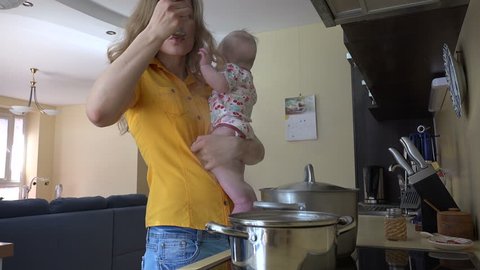 Young woman in yellow holding baby girl in arms, preparing food together in kitchen. Mother taste and salt food in steamy pot. Static shot. 4K UHD video clip.