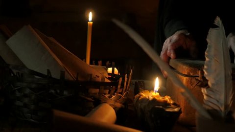 Dark Room in an old log hut of the 11th century. Candlelight. Writing Tools on Table, Old man, monk, chronicler, dressed in black robe, in the hood. Nestor the Chronicler. Man writes with a quill pen