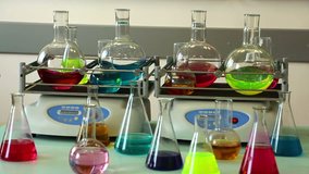 Laboratory shakers agitate eight round glass flasks filled with color solutions. Other glass wares stand in front. Focus on the shakers. Diagonal aspect angle. No camera movement.