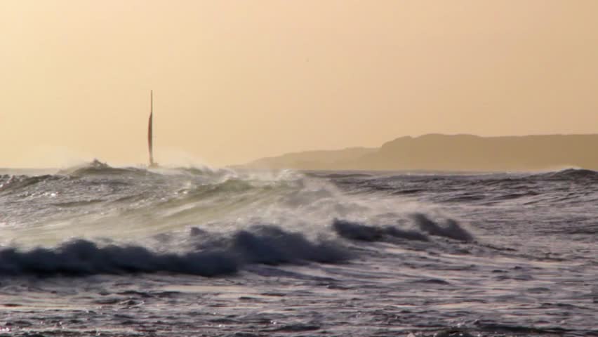 Surfers wait to catch a wave with a sailboat in the background