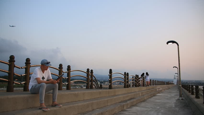 Man sitting at the bank and using smart phone and two young girls taking photos against airplane flying in sunset sky