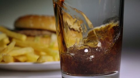 Cola being poured in glass, hamburger and potato chips on background,  slow motion, fast food, junk food concept.