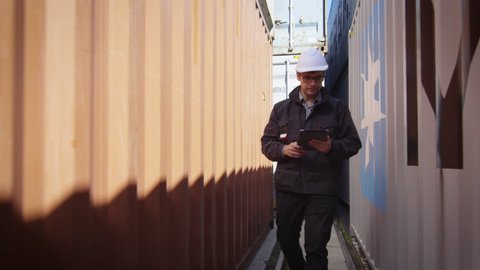 Worker in Hard Hat is Walking with Tablet PC between Containers in Logistic Center. Shot on RED Cinema Camera in 4K (UHD).