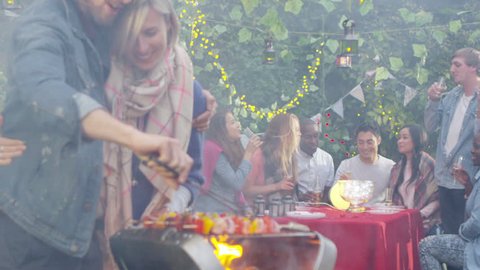 Happy couple share a kiss at bbq while friends socialize in the background. Shot on RED Epic.