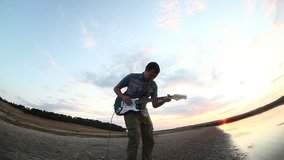 guitarist musician man playing solo on electric guitar by river at sunset sky  clouds 