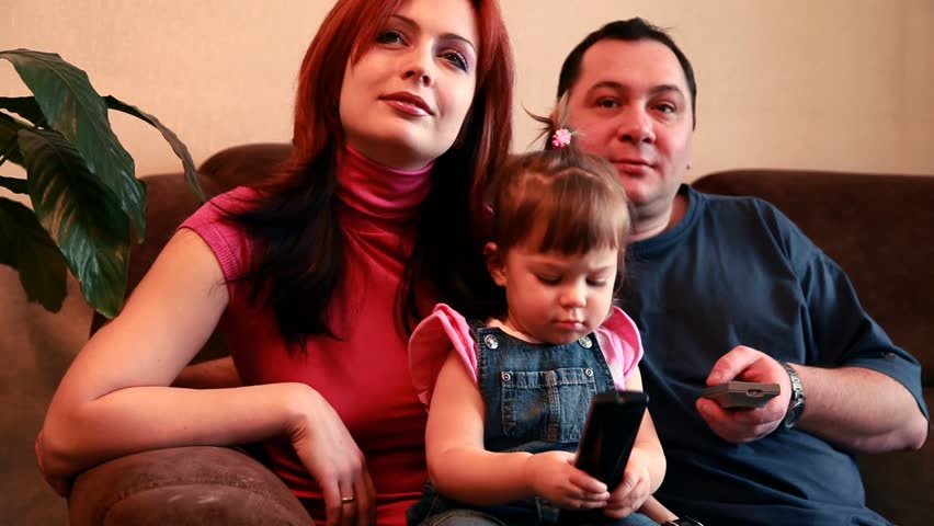 Parents and their baby girl are sitting on the sofa and watching TV with remotes in hands 
