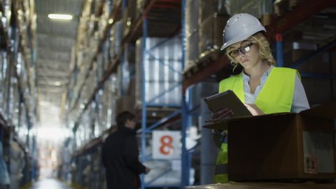 Female Manager of Logistics Warehouse is Working on Tablet PC. Wears a White Hard Hat. Shot on RED Cinema Camera in 4K (UHD).