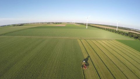 Aerial drone flying over a farmer in tractor working on grassland mixing mowing turning fresh grass in landscape with wind turbines windmills providing green energy renewable energy bird eye heli 4k