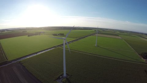 Drone aerial footage of wind turbine windmills during sundown sunset summer creating green energy renewable energy in background green grass land blue sky and sun helicopter bird eye cockpit view 4k
