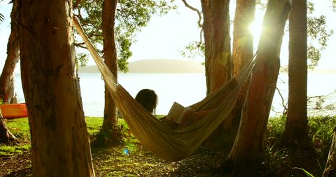 Woman relaxes reading book in hammock, swaying gently, in wood on beach at sunset. Sun breaks through trees