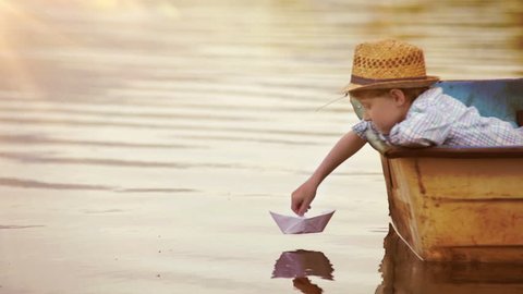 Little boy sitting in the real boat sets his paper ship off to sail
