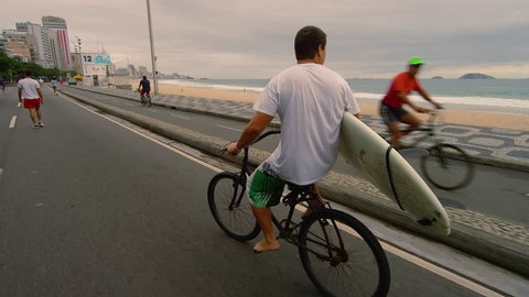 RIO DE JANEIRO - CIRCA JUNE 2013: Slow motion dolly shot of a man carrying a surfboard while riding his bike along the street near Ipanema Beach in Rio de Janeiro, Brazil. Adlı Haber Amaçlı Stok Video